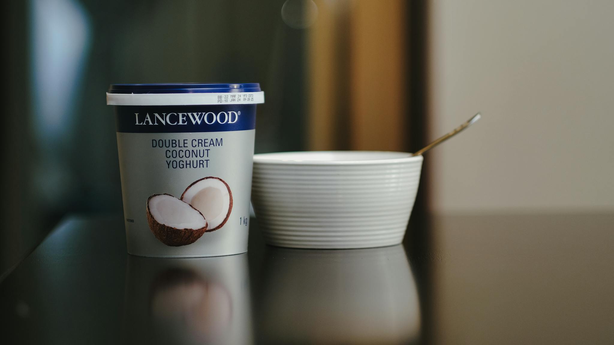 Lancewood double cream coconut yoghurt with a spoon and bowl on a reflective surface.