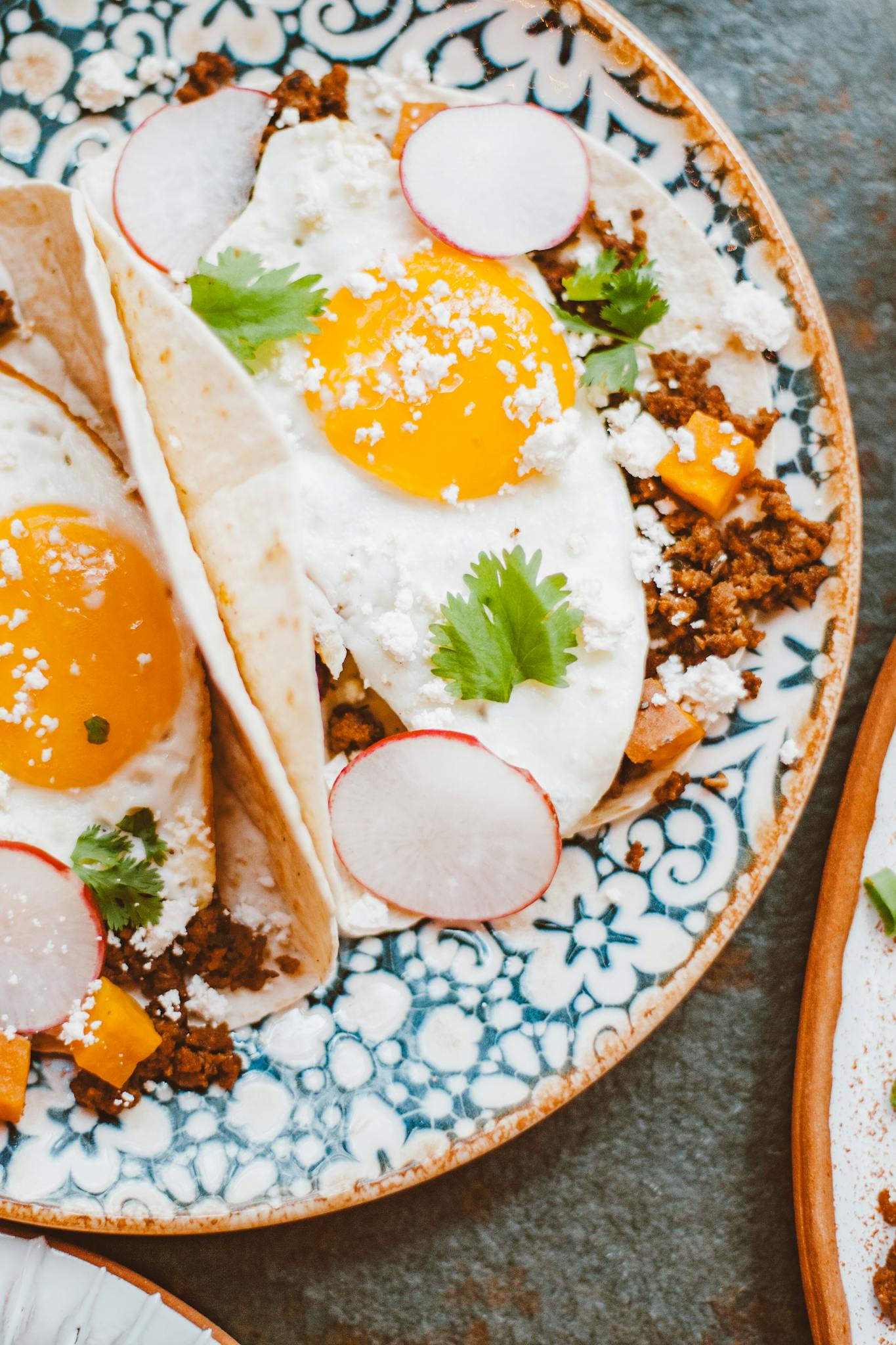 Top view of breakfast tacos with sunny-side-up eggs, radishes, and cilantro on a decorative plate.